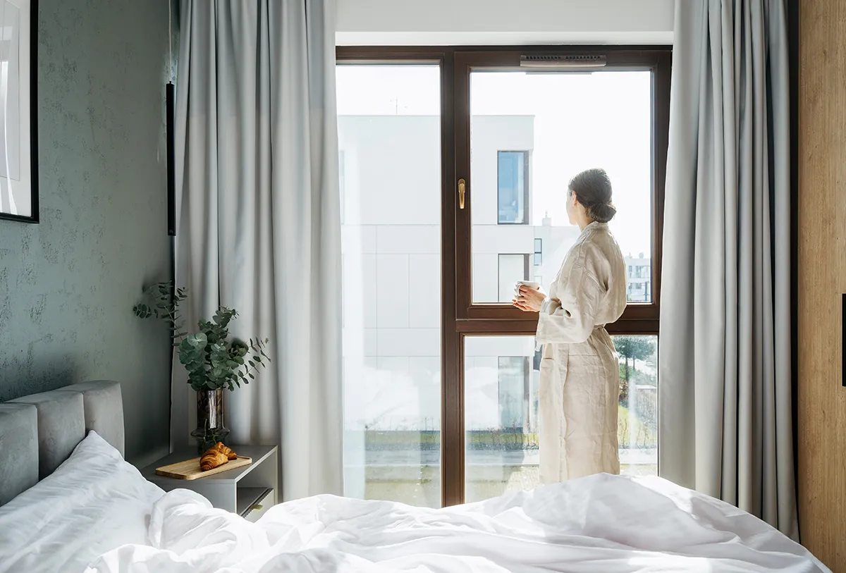 Kobieta w hotelowym pokoju stojąca przy oknie – symbol komfortu i bezpieczeństwa zapewnianego przez drzwi przeciwpożarowe